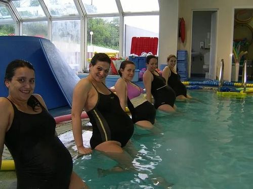 Mujeres embarazadas en una piscina, sonriendo, haciendo ejercicios acuáticos en una habitación luminosa.