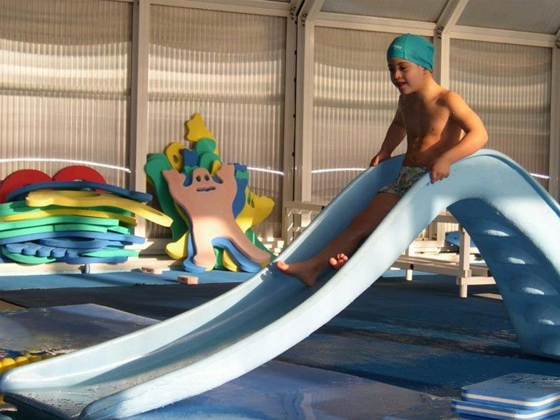 Un niño se desliza en una piscina azul. Lleva gorro de baño y bañador. La luz se refleja en el agua.