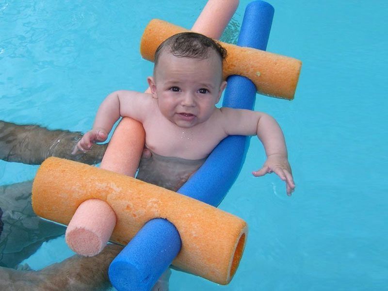 Bebé en la piscina, rodeado de flotadores naranjas y azules. Ojos alerta, sostenidos por manos invisibles.