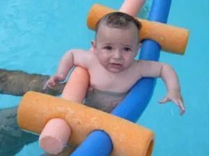 Bebé en una piscina, sostenido por fideos de espuma de colores, con aspecto ligeramente aprensivo.