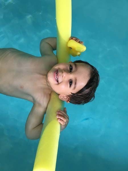 Niño sonriendo en una piscina, sosteniendo un fideo amarillo, con un pequeño patito de goma.