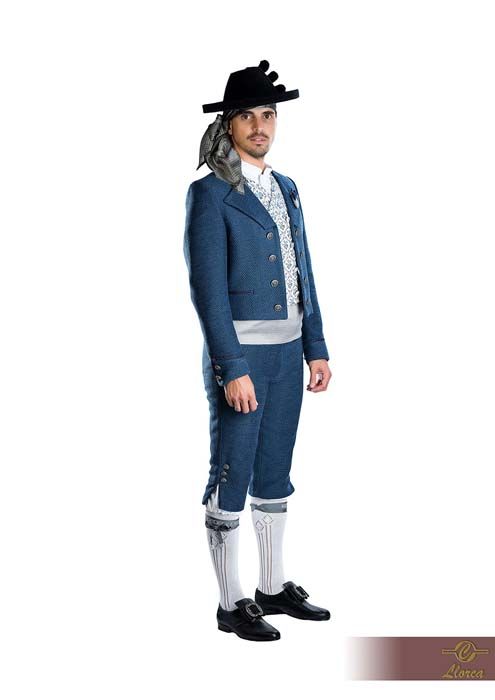 Una persona que viste un traje folclórico español tradicional de color azul, que incluye un chaleco estampado, pantalones hasta la rodilla y un sombrero oscuro.