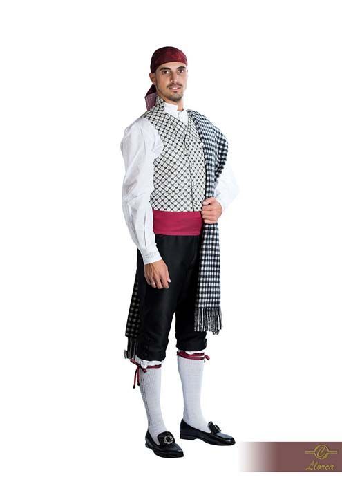 Una persona vestida con un traje folclórico tradicional español: camisa blanca, chaleco estampado, pantalones oscuros, fajín y chal a cuadros.