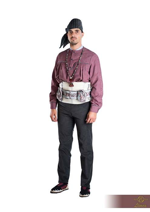 Una persona que viste una camisa abotonada de color malva, pantalones oscuros, un cinturón ancho blanco estampado, un pañuelo oscuro en la cabeza y un collar de cuentas.