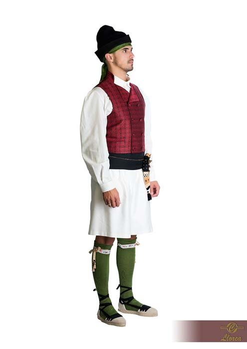 Una persona vestida con un traje tradicional compuesto por un chaleco rojo, camisa blanca, falda escocesa, calcetines verdes y gorro negro, de pie de lado.