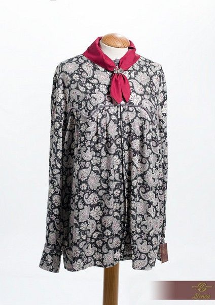 Una camisa de manga larga, de color gris y negro con estampado de cachemir, exhibida en un maniquí, complementada con una bufanda rosa vibrante al cuello.