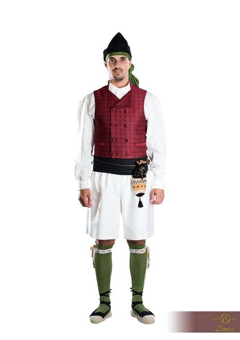 Un hombre con traje tradicional compuesto por un chaleco rojo estampado, camisa blanca, pantalones cortos blancos, calcetines verdes y gorra negra.