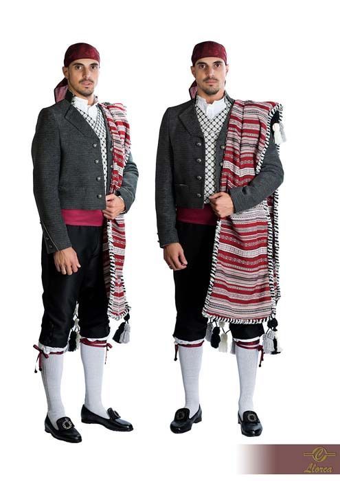 Dos figuras con trajes tradicionales sardos, que visten chaquetas oscuras, pantalones negros, fajas rojas en la cintura y chales a rayas.