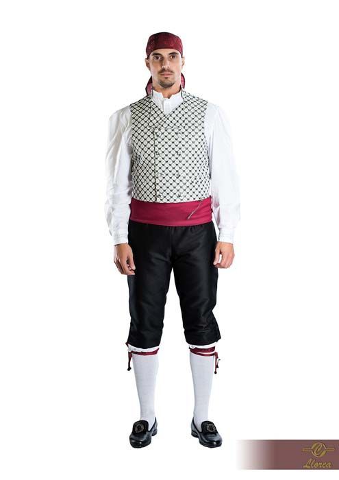Un hombre que viste un traje tradicional con chaleco estampado, camisa blanca, pantalones negros, faja roja y turbante.