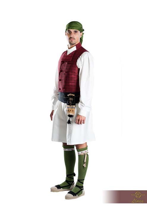 Una persona con el traje tradicional mediterráneo: pañuelo verde en la cabeza, camisa blanca, chaleco granate, falda escocesa blanca y polainas verdes.