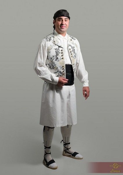Una persona viste un atuendo tradicional blanco que incluye un chaleco estampado, una faja oscura en la cintura, una diadema y zapatos con cordones.