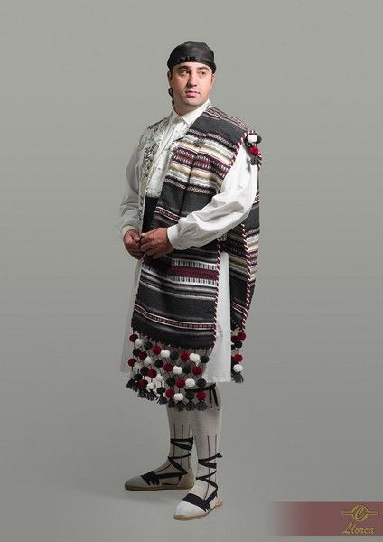 Una persona aparece vestida con ropa tradicional: una camisa blanca, un manto de lana estampado con pompones, polainas ligeras y sandalias.