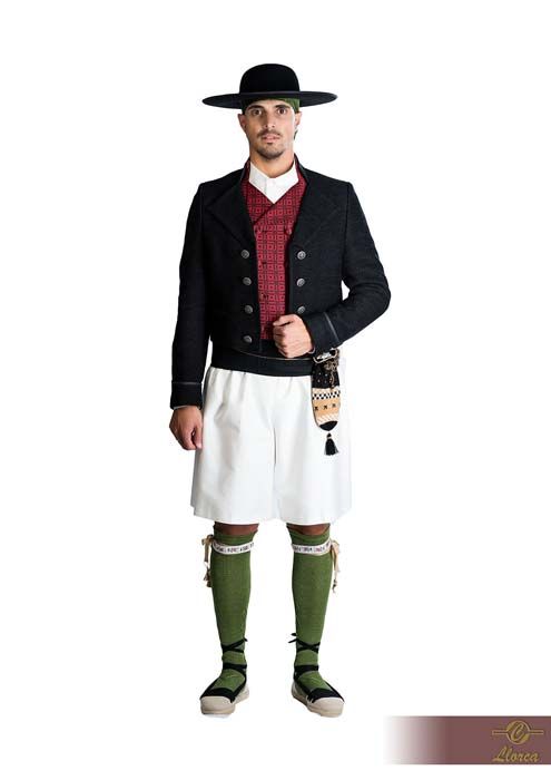 Una persona que lleva un sombrero negro de ala ancha, una chaqueta oscura, pantalones cortos blancos, un chaleco rojo estampado, calcetines verdes y zapatos con cordones.