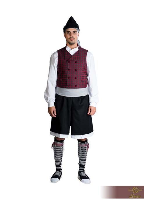 Una persona que viste un traje folclórico tradicional que consiste en un chaleco oscuro estampado, una camisa blanca, pantalones cortos negros y calcetines a rayas.