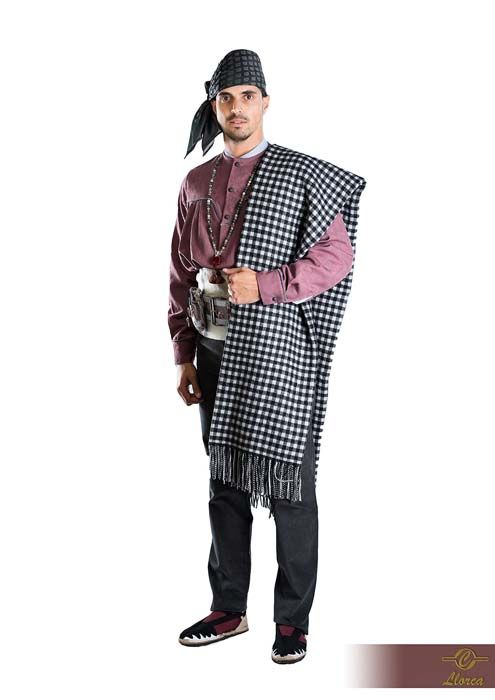 Una persona vestida con el atuendo tradicional cretense: camisa granate, pantalones oscuros, pañuelo estampado en la cabeza y chal con flecos.