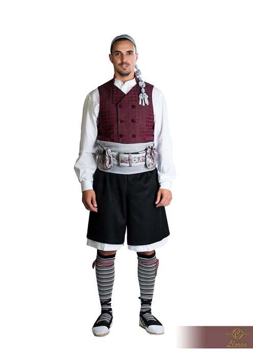 Persona con atuendo tradicional: chaleco granate, camisa blanca, pantalones cortos negros, calcetines a rayas y una ancha faja gris sobre fondo blanco.