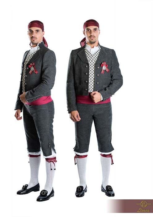Dos personas vestidas con trajes tradicionales de color gris oscuro, pañuelos rojos en la cabeza, chalecos estampados y medias blancas sobre un fondo blanco.