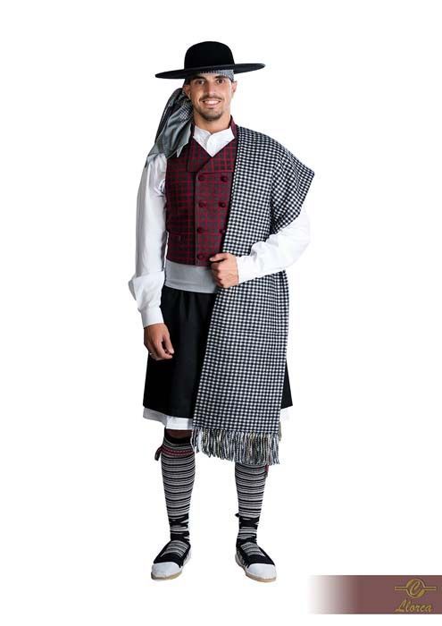 Una persona sonriente que viste un atuendo tradicional compuesto por sombrero, chaleco color burdeos, camisa blanca, falda escocesa negra y chal estampado.