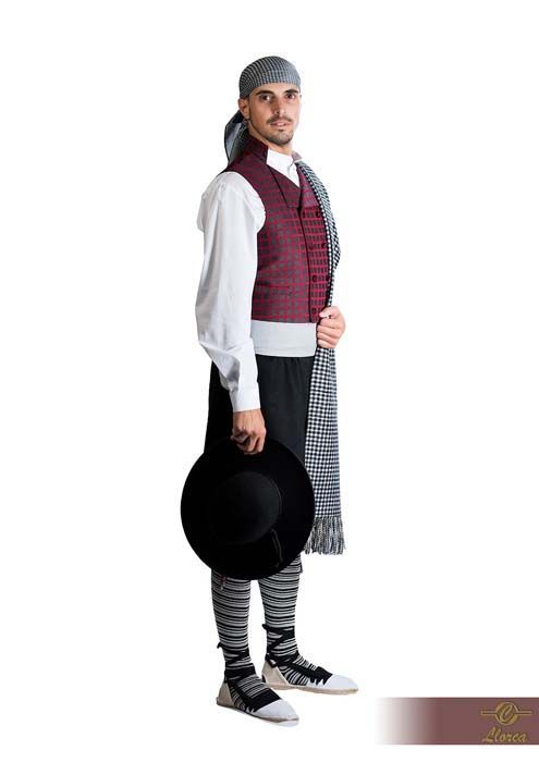 Una persona que viste ropa tradicional, que incluye un chaleco granate, una camisa blanca, un pañuelo estampado en la cabeza y mallas a rayas.