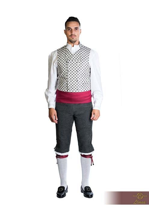 Una persona vestida con un atuendo tradicional: camisa blanca, chaleco cruzado estampado, fajín rosa, pantalones grises y calcetines.