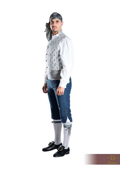 Una persona luciendo un traje folclórico tradicional y formal que consiste en un chaleco estampado, una camisa blanca, pantalones azules y calcetines.