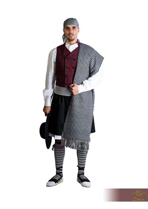 Una persona vestida con un traje folclórico tradicional español: camisa blanca, chaleco color burdeos, falda negra, chal estampado y sombrero.