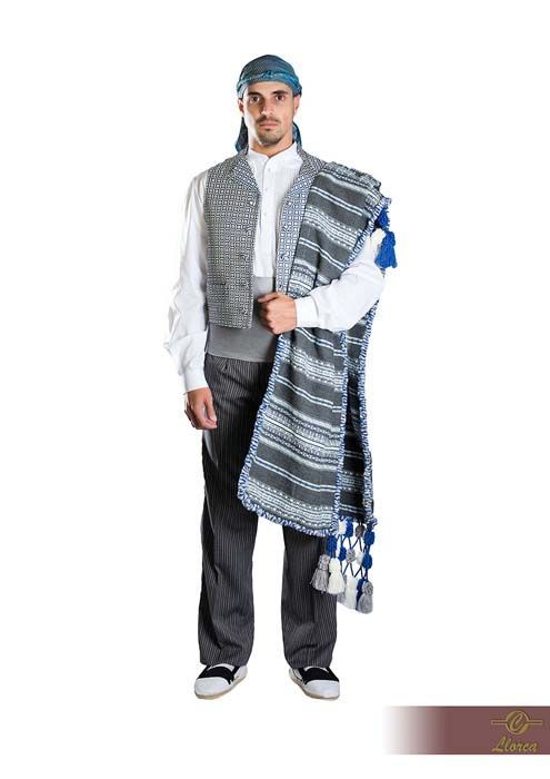 Una persona que viste un atuendo tradicional compuesto por una camisa blanca, un chaleco gris, pantalones oscuros, un pañuelo en la cabeza y un chal azul estampado.
