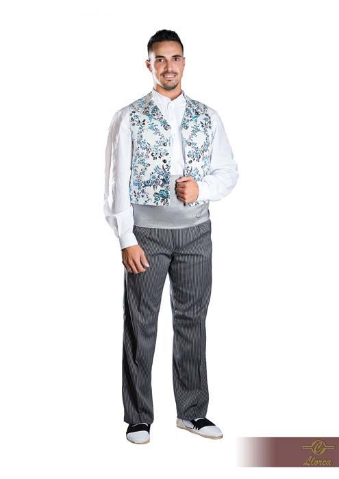 Una persona lleva una camisa blanca, un chaleco azul claro con estampado floral, una faja ancha gris y pantalones oscuros texturizados.
