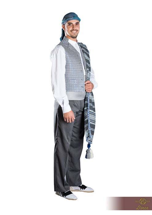 Una persona sonriente que lleva un pañuelo estampado en la cabeza, una camisa blanca de manga larga, un chaleco plateado, pantalones oscuros y una faja azul.