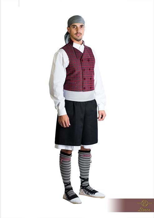 La persona viste atuendo folclórico tradicional: una camisa blanca, un chaleco granate, pantalones cortos negros, calcetines altos a rayas y un pañuelo en la cabeza.