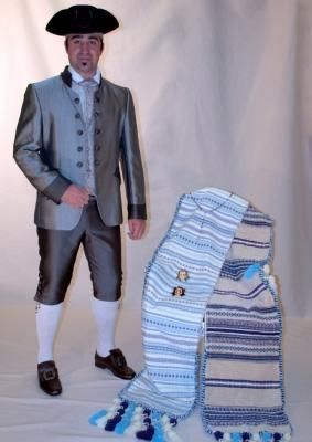 Una persona vestida con un traje gris de época y un sombrero tricornio se encuentra de pie junto a un chal tejido, doblado, de color azul, blanco y marrón.