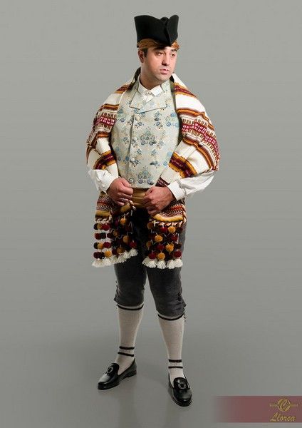 Un hombre viste ropa folclórica tradicional que incluye un chaleco estampado, una estola tejida a rayas, pantalones hasta la rodilla y un sombrero.