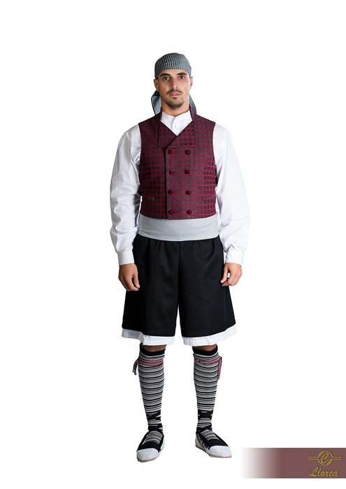 Un hombre con un traje tradicional que incluye un pañuelo estampado en la cabeza, un chaleco cruzado color burdeos, pantalones cortos negros y mallas.