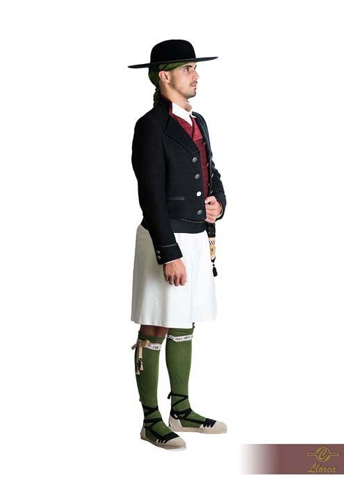 Una persona aparece de pie, vestida con un traje folclórico tradicional vasco: chaqueta oscura, falda blanca, calcetines verdes y sombrero de ala ancha.