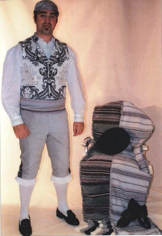 Una persona vestida con un traje folclórico tradicional español está de pie junto a una silla cubierta con un sarape a rayas y un sombrero negro.