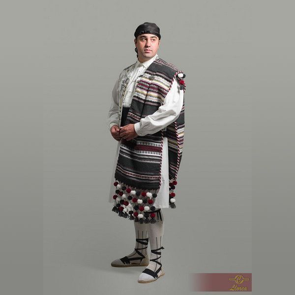 Una persona luce una prenda tradicional cretense que consiste en una capa de lana a rayas con pompones, una túnica blanca y calzado con cordones.