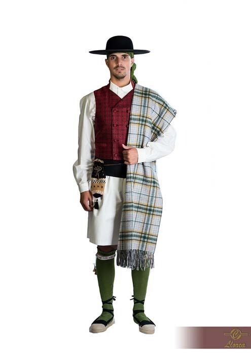 Una persona que lleva un sombrero de ala ancha, un chaleco granate, una camisa blanca, un chal estampado de color claro y vendas verdes para las piernas.