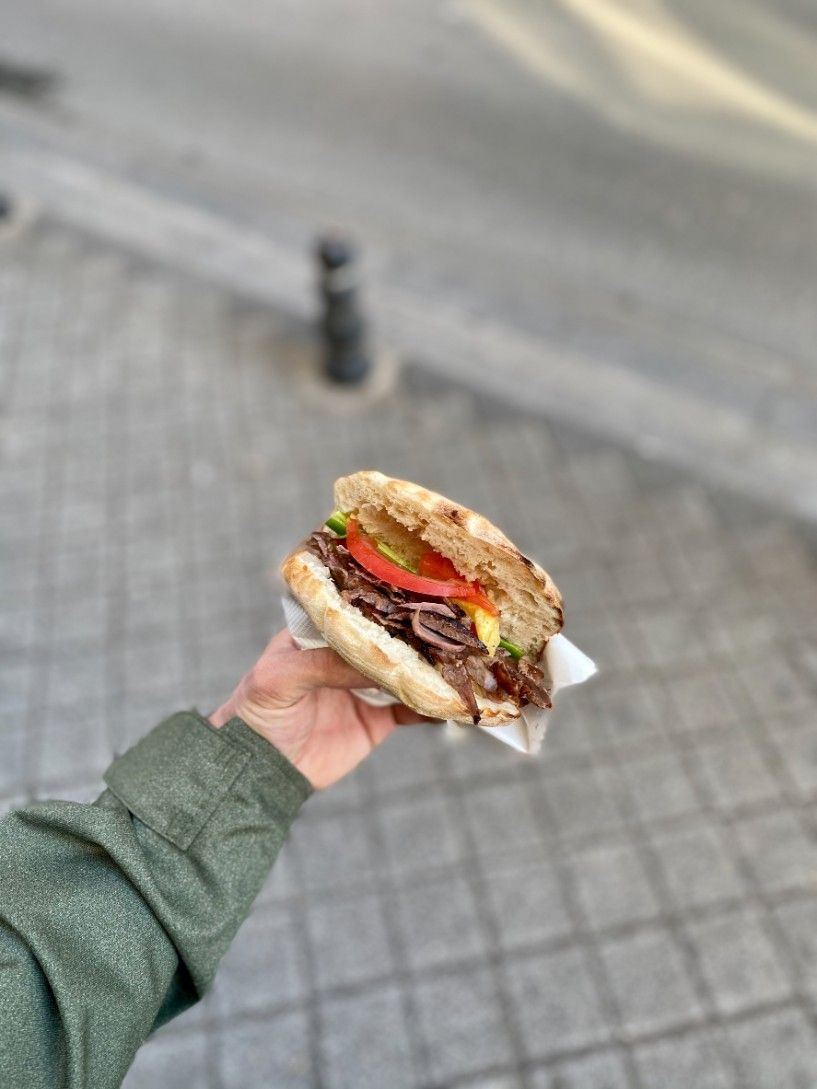 Mano sosteniendo un sándwich con carne, tomate y lechuga; entorno al aire libre, suelo pavimentado.