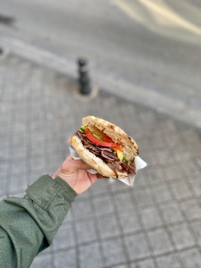 Mano sosteniendo un sándwich con carne, tomate y lechuga; entorno al aire libre, suelo pavimentado.