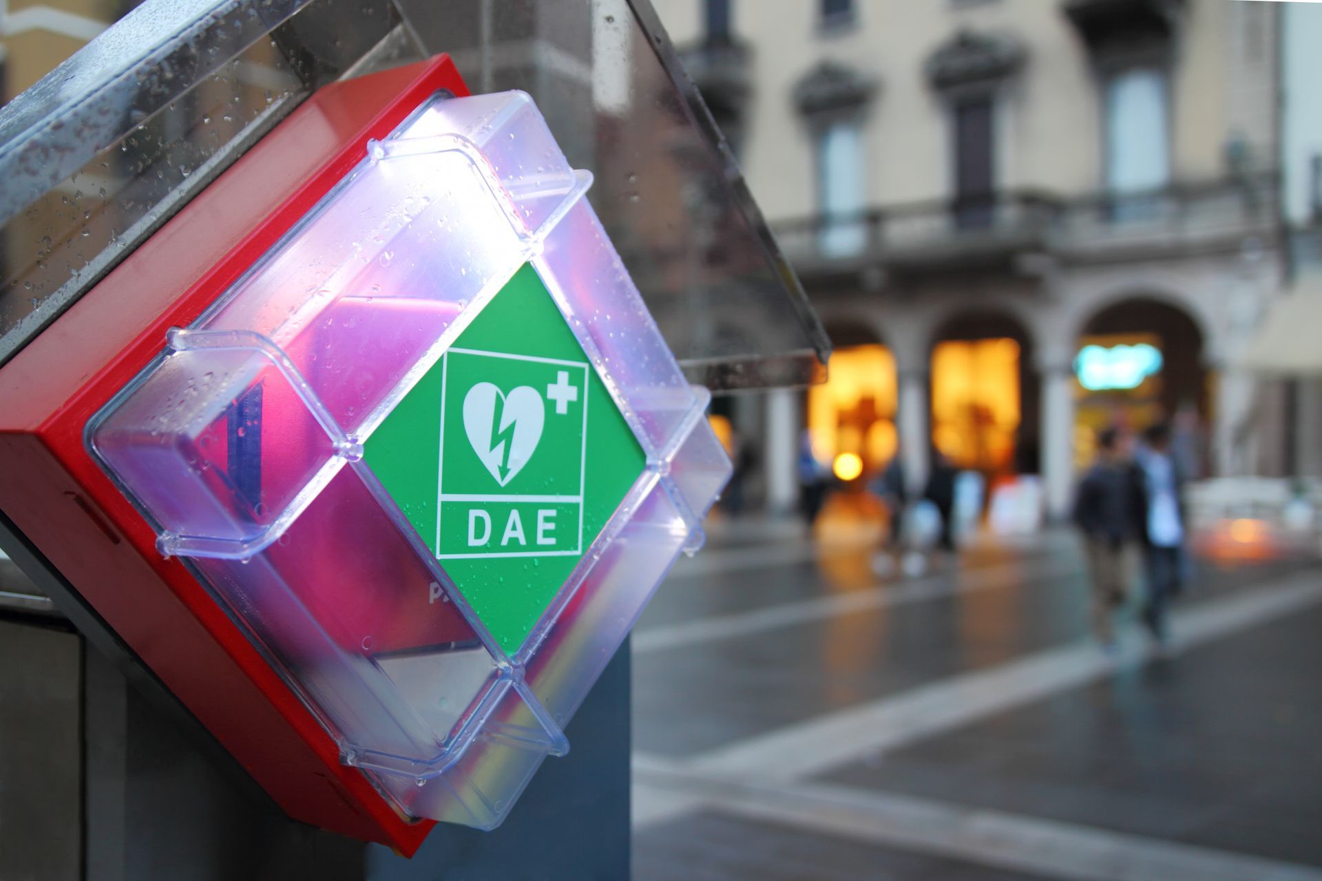 DEA (défibrillateur) dans une boîte rouge avec un symbole de cœur vert ; à l'extérieur, personnes et bâtiments flous en arrière-plan.