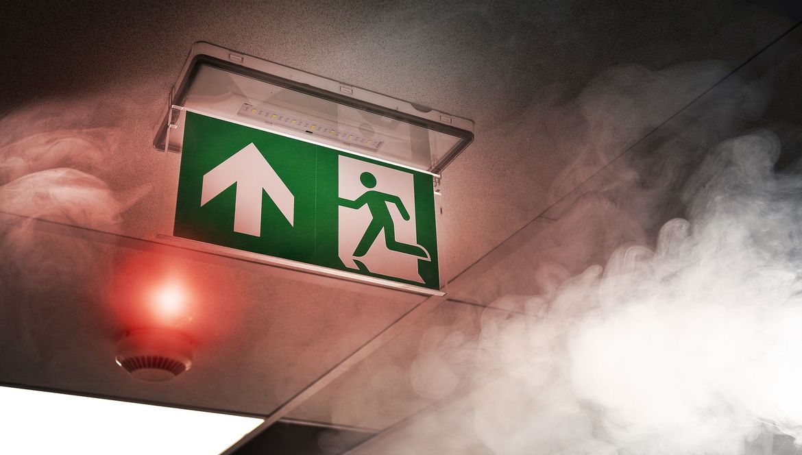 Panneau de sortie de secours, vert, au-dessus d'une personne qui court, avec de la fumée et une alarme rouge en arrière-plan.