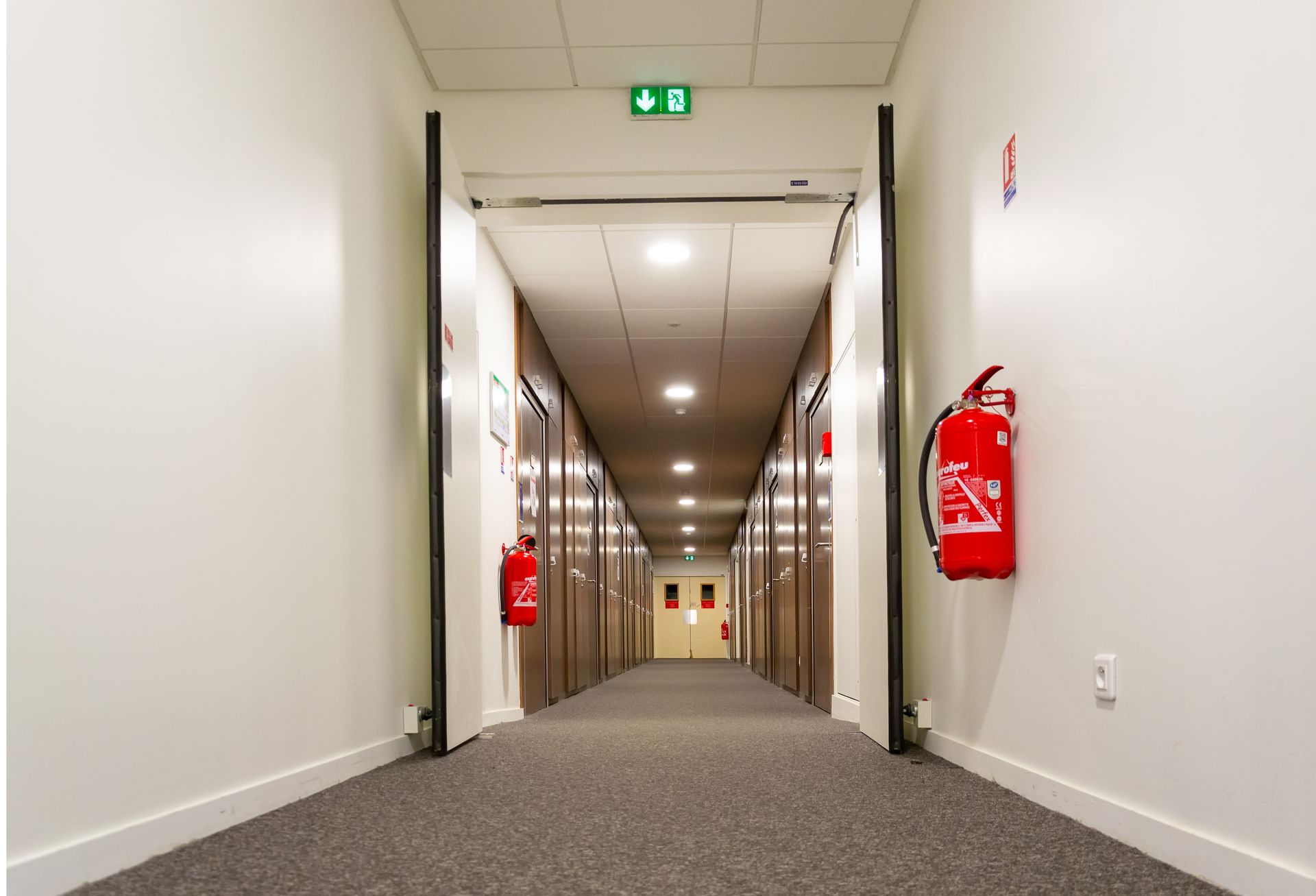 Long couloir d'hôtel avec portes, extincteurs et panneau de sortie.