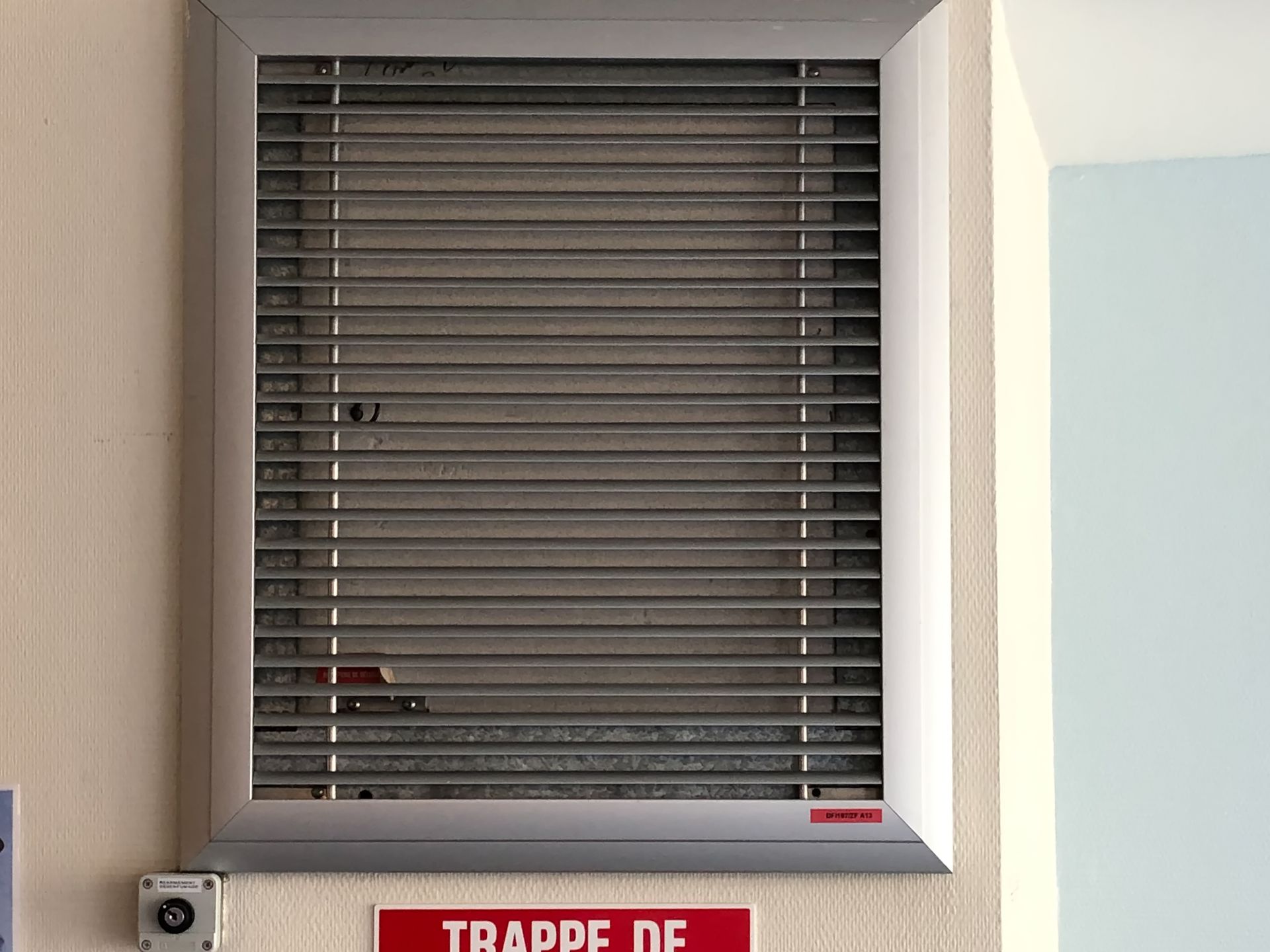 Grille de ventilation argentée à persiennes encastrée dans un mur beige.