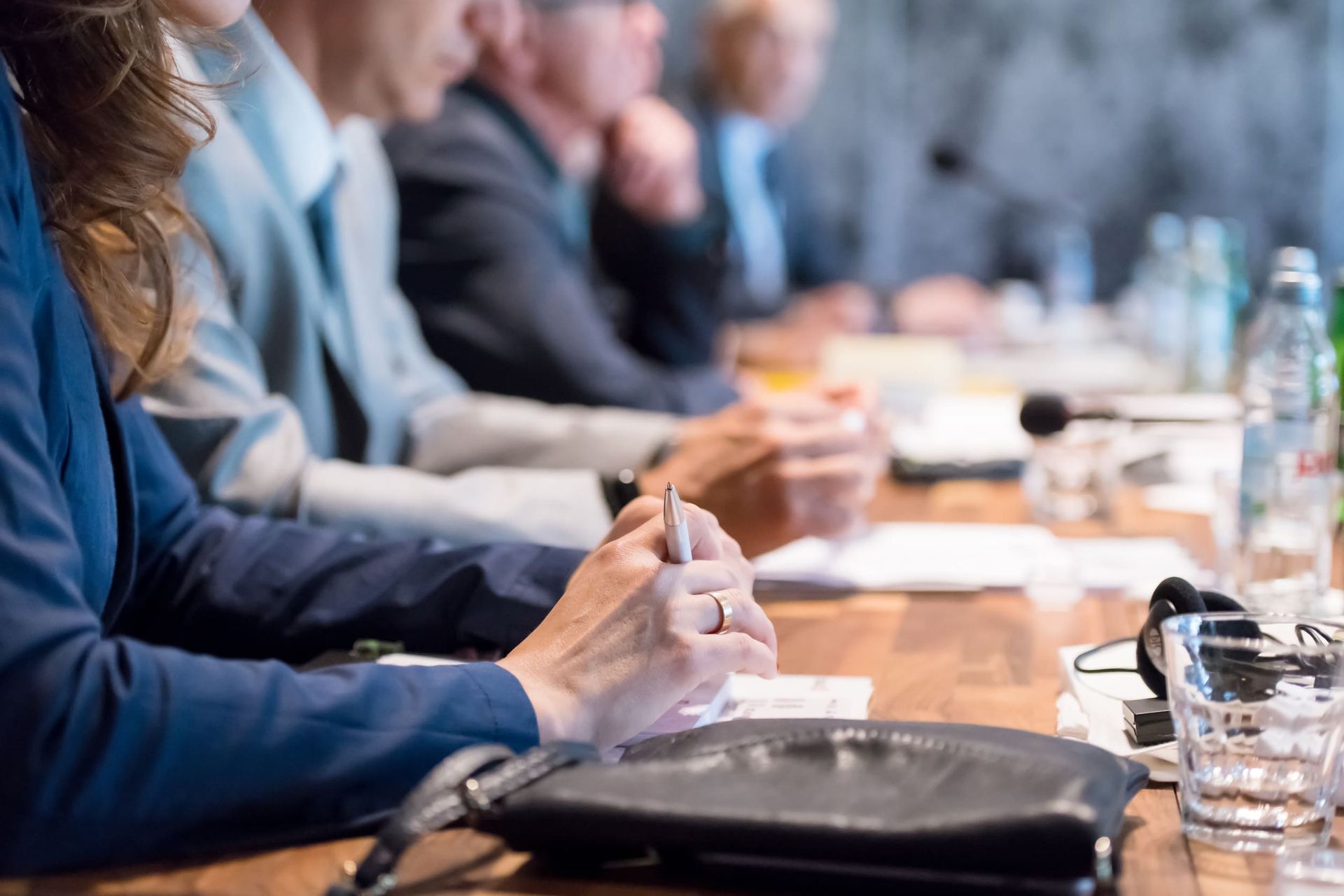 Personas sentadas a una mesa en una reunión, con las manos sosteniendo bolígrafos, micrófonos y botellas de agua.