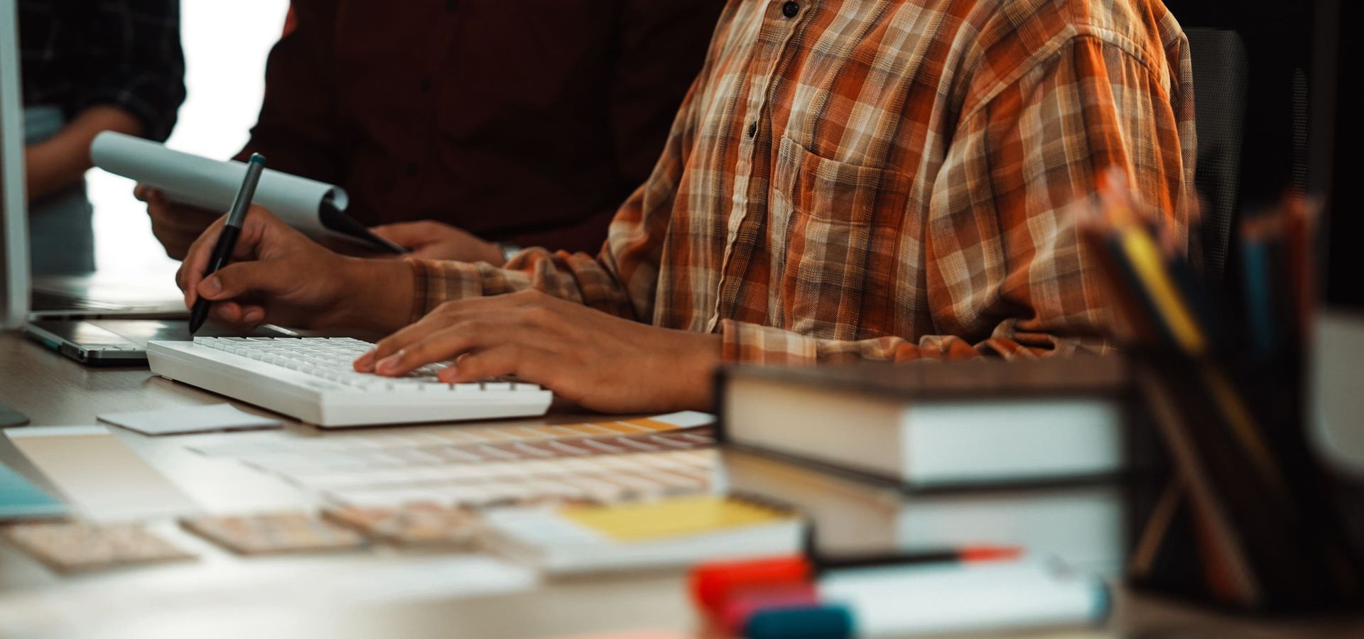 Persona trabajando en un ordenador y escribiendo en papel. Escritorio con libros y material de oficina.