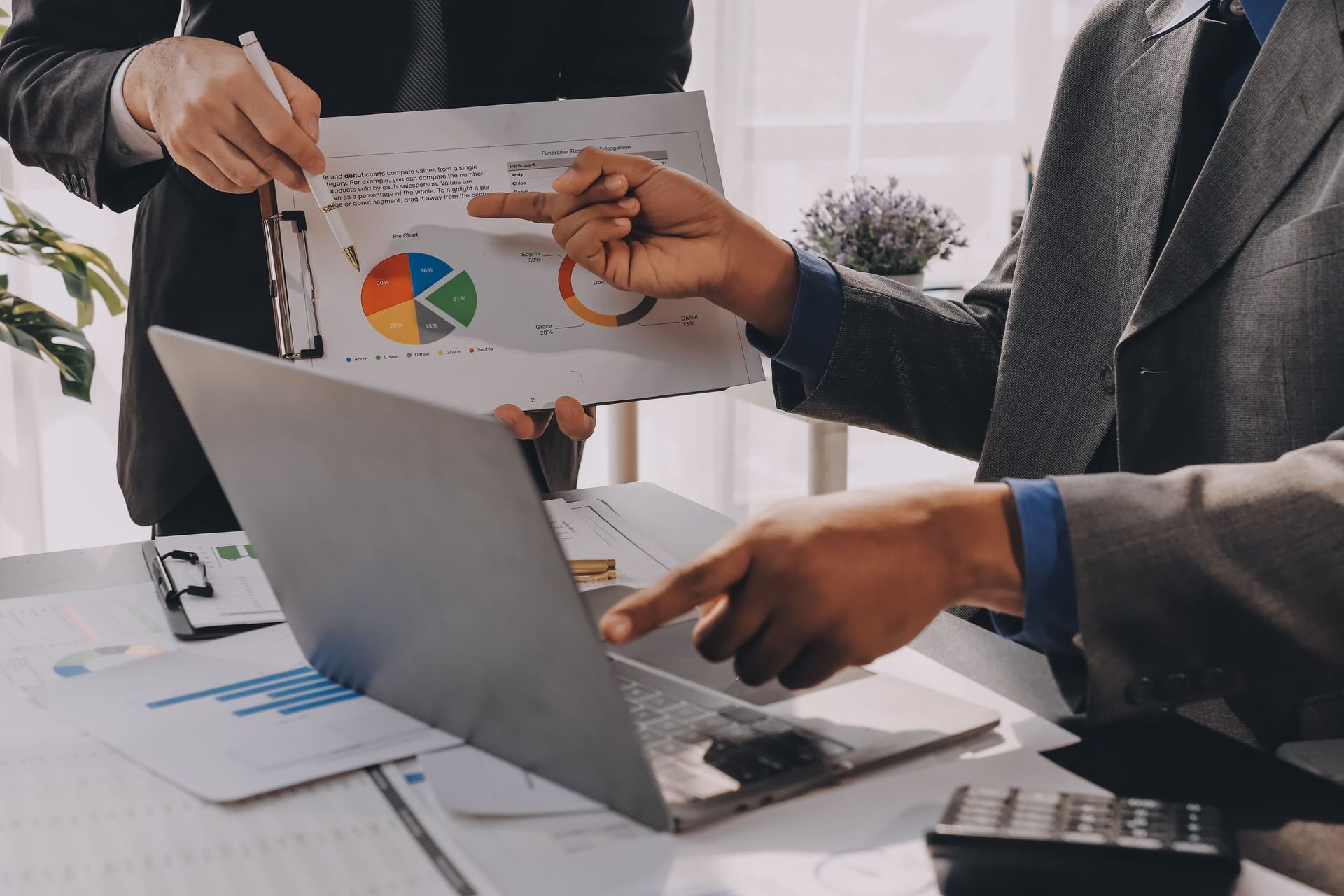 Dos personas revisan gráficos y una computadora portátil en un escritorio. Una señala un gráfico circular, mientras que la otra señala la computadora portátil.