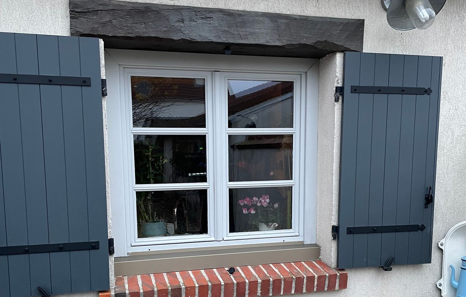Fenêtre avec volets bleus ouverts, cadre blanc, appui de fenêtre en briques et poutre en bois sombre au-dessus.