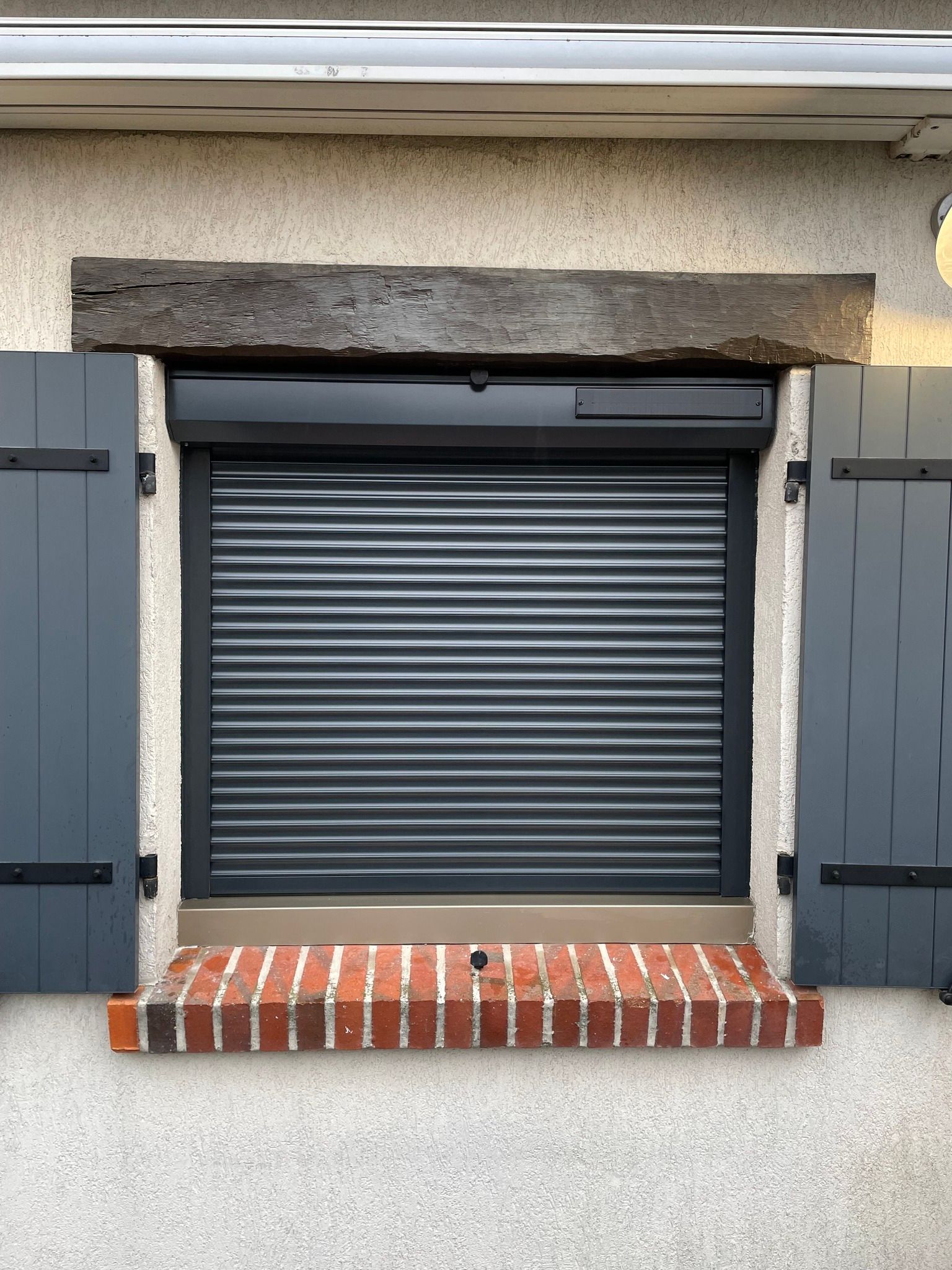 Un volet roulant gris foncé recouvre une fenêtre à volets gris, posée sur un appui en briques.