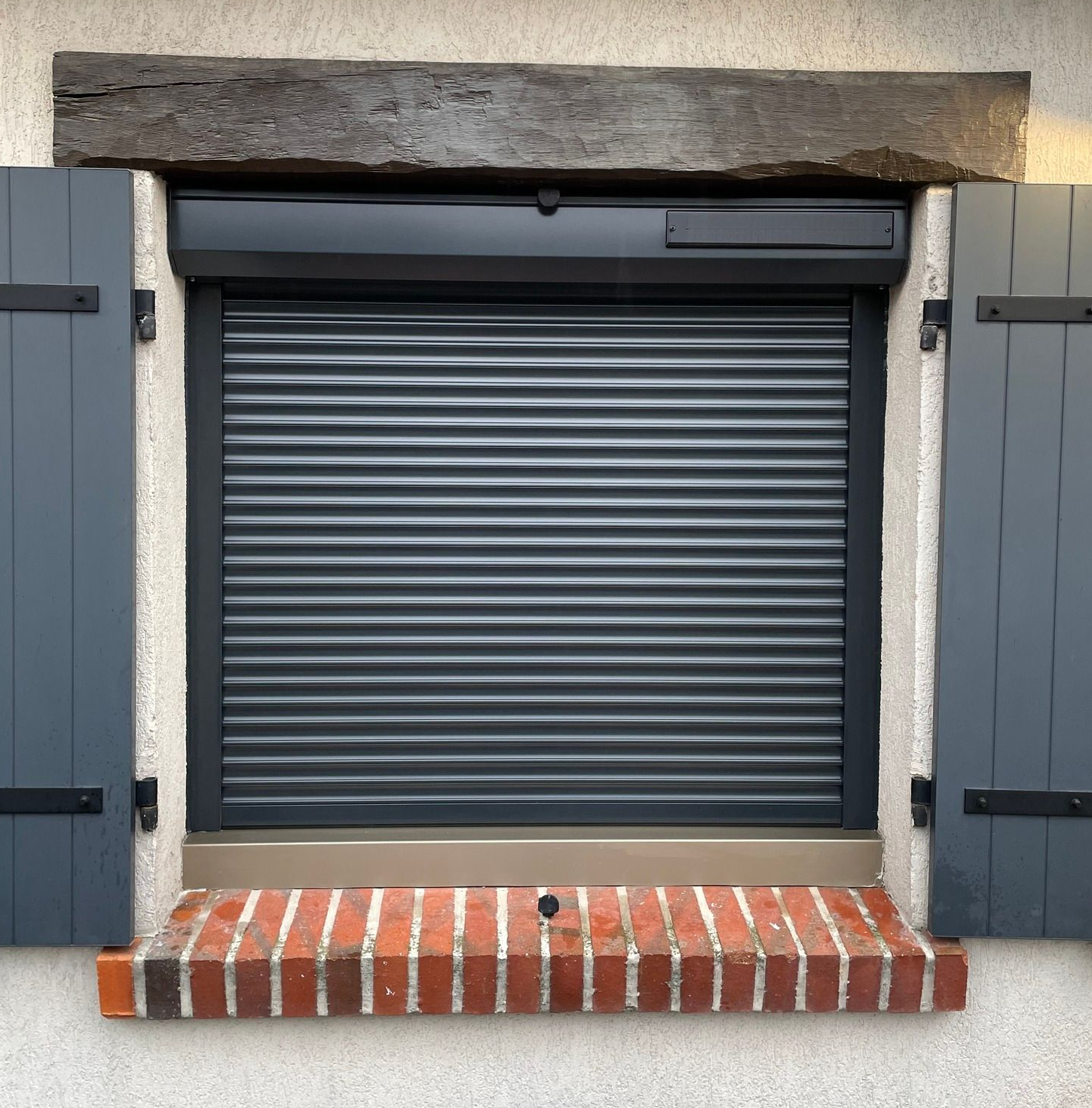 Fenêtre grise avec volet roulant fermé, appui de fenêtre en brique et volets gris. Encastrée dans un mur en pierre.