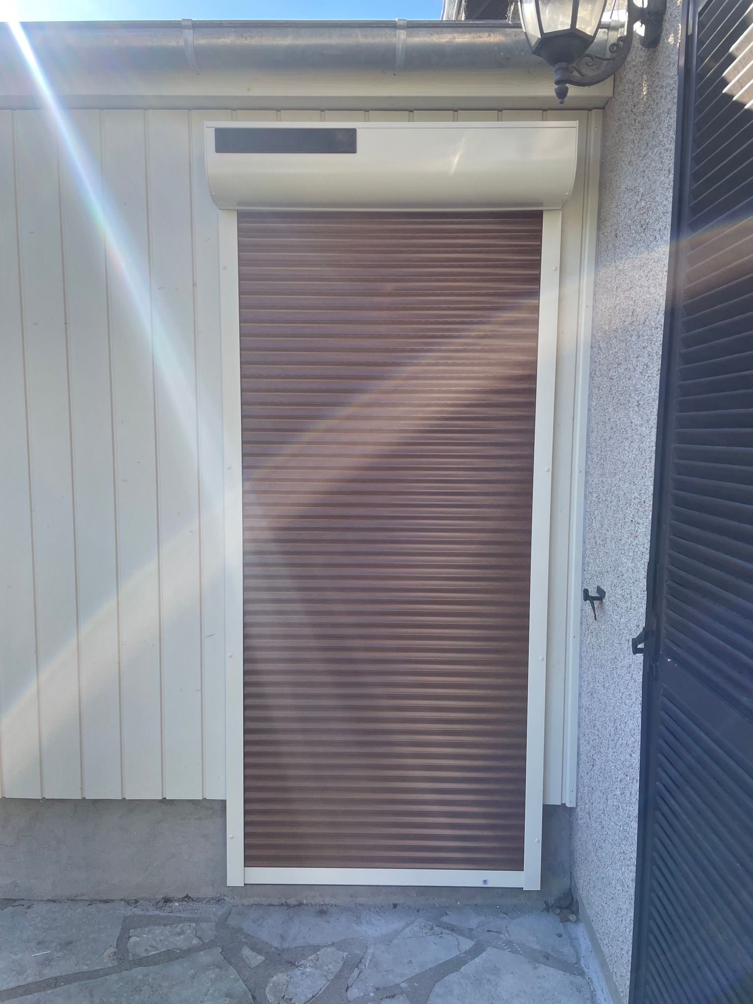Volet roulant marron sur un bâtiment beige, baigné par les reflets du soleil.